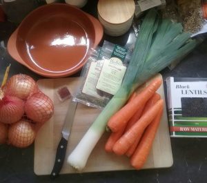 chopping board with arrangment of carrots, leek, onion, spices, lentils, and an earthenware dish.